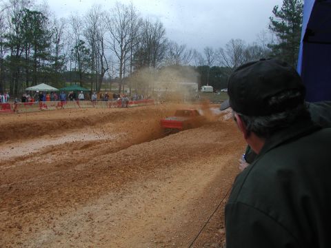 1985 Ford Bronco - A red truck navigates a muddy track during a competition, with spectators observing the event.