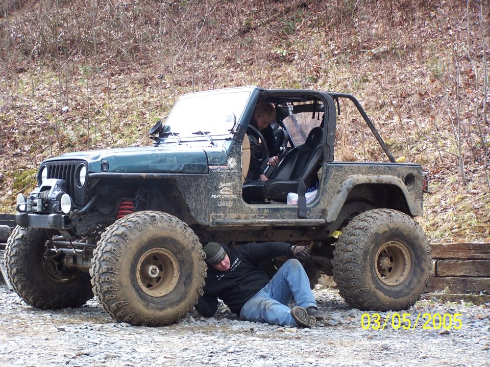 Guy working under a lifted Jeep Wrangler with big tires. Another person is in the driver's seat