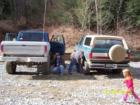 1979 Ford F-150 - Two men inspect tires on a green Ford Bronco and a blue and white pickup truck in a wooded area.