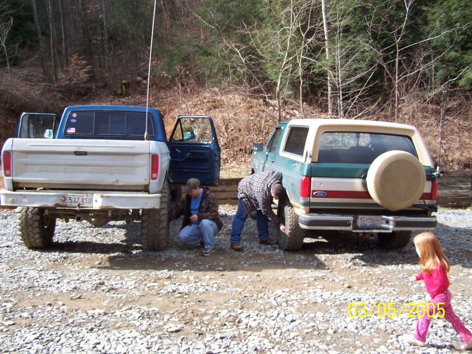 Green Ford Bronco with a spare tire on the back, parked next to a blue and white pickup truck