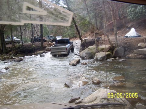 1979 Ford F-150 - A silver 1979 Ford F-150 navigates a rocky stream with other vehicles and a tent visible in the background.