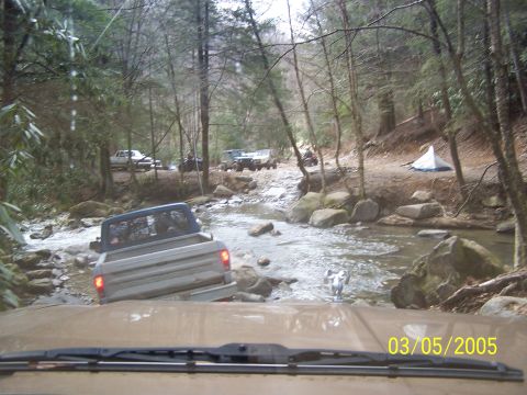 1979 Ford F-150 - A group of trucks navigates a rocky stream bed in a forest setting, with a tent visible on the side.