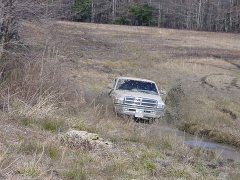 1989 Ford Bronco - A light-colored pickup truck navigates a muddy terrain, kicking up dirt and water.