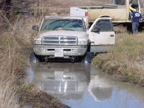 1989 Ford Bronco - A muddy pickup truck is stuck in water, with another truck visible behind it and people standing nearby.