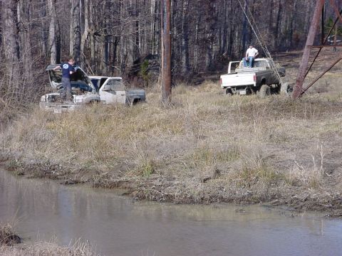 1989 Ford Bronco - Two trucks are stuck in a muddy, wooded area, with one vehicle's hood open as people attend to it.