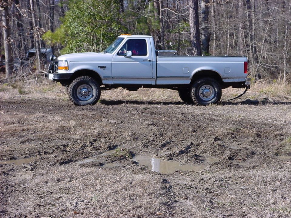White pickup truck with a toolbox in the bed, parked in a muddy area with trees in the background