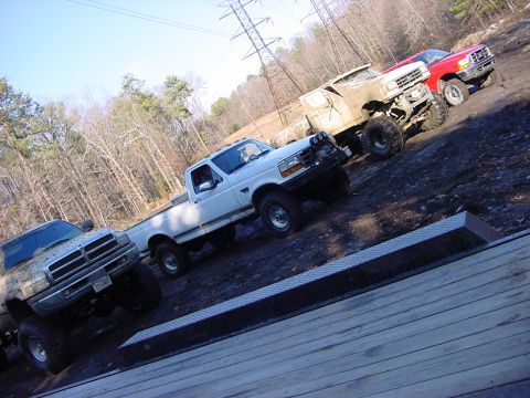 1989 Ford Bronco - Several modified trucks are parked on a muddy area with trees and power lines visible.