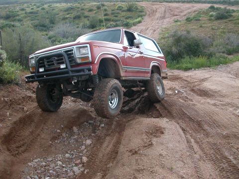 1984 Ford Bronco - This lifted Ford Bronco is shown off-roading on a dirt path with hills in the background.