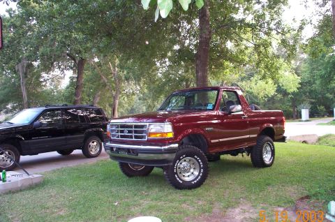 1996 Ford Bronco - Lifted 1990s maroon Ford full-size Bronco on chrome wheels and off-road tires, parked under trees next to a dark Jeep.