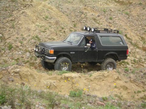 1988 Ford Bronco - A black Ford Bronco navigates a challenging off-road trail with a driver at the wheel.