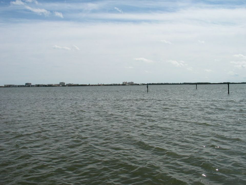 Water with distant buildings under a cloudy sky. Three posts are visible in the water