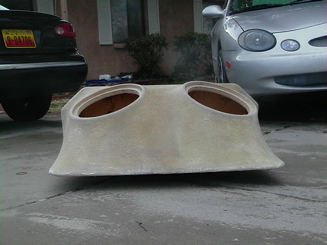 Raw fiberglass dual-subwoofer box on driveway with two cars behind it