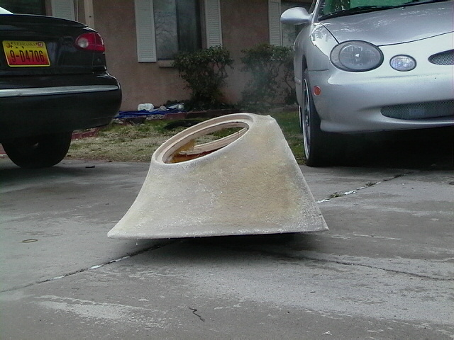 Unpainted wedge-shaped fiberglass shell on a driveway between two cars