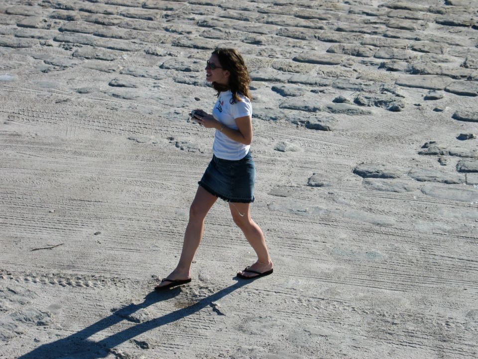 Woman in white t-shirt and denim skirt walks on a textured paved area. Supermotors.net logo in lower right