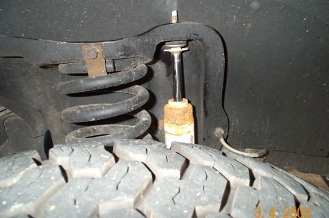 1996 Ford Bronco - Close-up under a wheel well showing a coil spring, a white-bodied shock absorber with a rust-stained upper body and exposed chrome shaft, t…