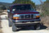 1996 Ford Bronco - Very small, low-resolution front view of a dark-colored full-size Ford SUV parked on a dirt or gravel area with green brush behind it.