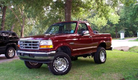 1996 Ford Bronco - maroon lifted Ford Bronco with chrome wheels and the top off, parked on grass in a wooded yard.