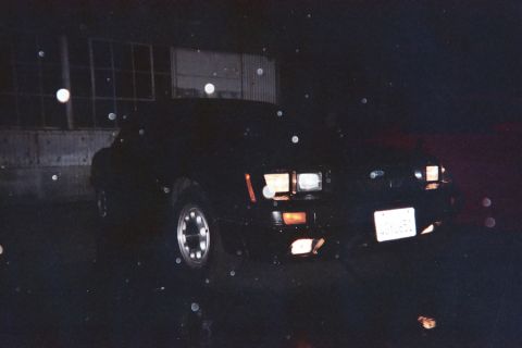 1985 Ford Mustang - A black 1985 Ford Mustang is parked outside a building at night, with its headlights illuminated.