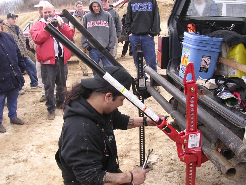 Man using a red Hi-Lift jack at the rear of an SUV as a crowd watches outdoors