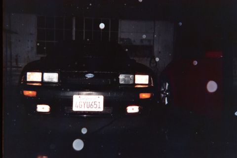 1985 Ford Mustang - Dark night front view of a black Fox-body Mustang with quad rectangular headlights, amber turn signals and lower fog lamps lit, a Ford oval…