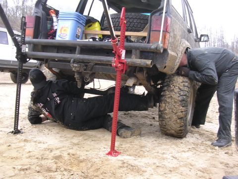 1995 Jeep Cherokee - Two individuals are performing maintenance on a muddy Jeep Cherokee, with a second vehicle partially visible in the background.