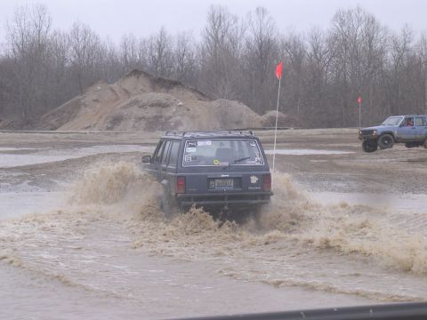 1995 Jeep Cherokee - A dark Jeep Cherokee navigates a muddy water obstacle, with another vehicle airborne in the background.