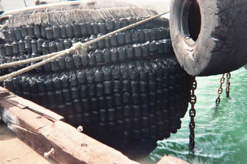 1990 Ford Bronco - Rows of black tires bound as marine fenders cover a vessel's hull at a dock, with chains and rope and green water below.