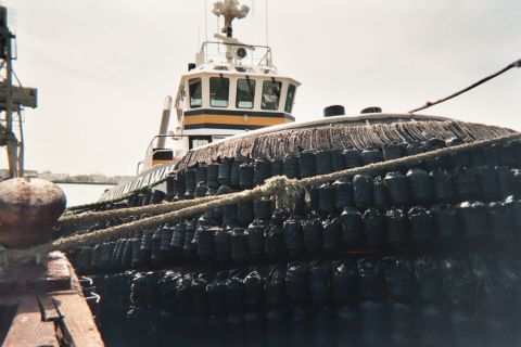 1990 Ford Bronco - tugboat with a white-and-yellow wheelhouse and tire-covered hull sits moored at a harbor pier.