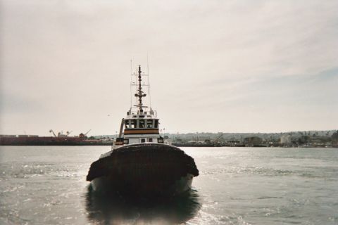 1990 Ford Bronco - tugboat seen head-on moves across sunlit water with cranes and a port shoreline in the hazy background.