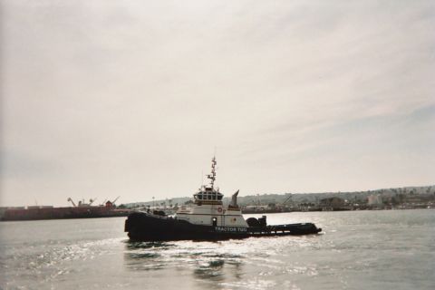1990 Ford Bronco - black-hulled tractor tug labeled 'Tractor Tug' cruises across a calm harbor with a hillside port behind it.