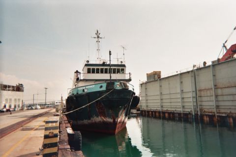 1990 Ford Bronco - weathered blue-green workboat with a tall wheelhouse is moored bow-out at a dock beside a rusty barge.