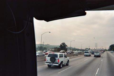 1990 Ford Bronco - white Ford Bronco with a spare tire on the tailgate travels a multi-lane freeway, viewed through a vehicle's side window.
