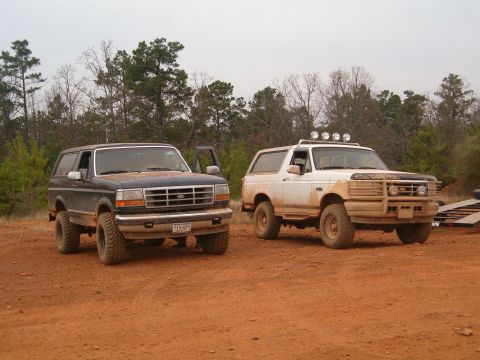 1996 Ford Bronco - Two Ford Broncos are parked on a dirt lot with trees in the background. The white Bronco is covered in mud.