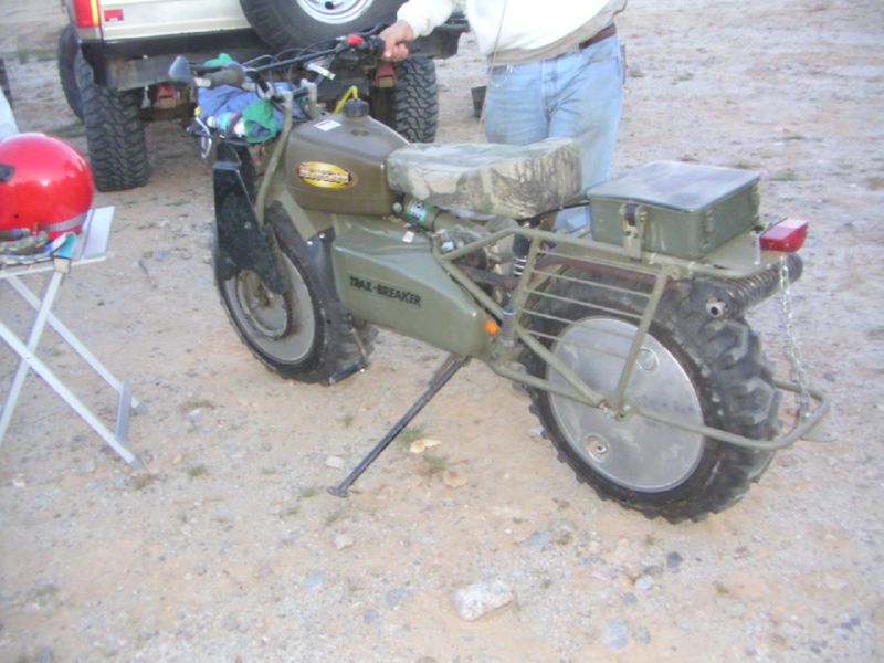 Olive-drab tracked minibike with camo seat parked on gravel by a 4x4