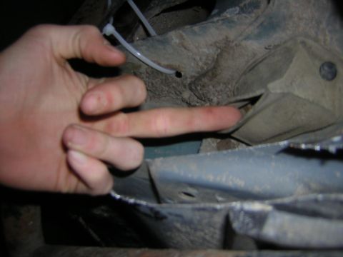 1995 Ford Bronco - Close-up of a hand pointing to a metal bracket on the underside of a vehicle, possibly for inspection or repair.