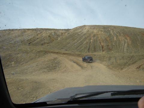 1991 Ford Bronco - Shot through a windshield across a dark dashboard and wiper, looking out at a dark full-size SUV with a rear-mounted spare tire climbing a…