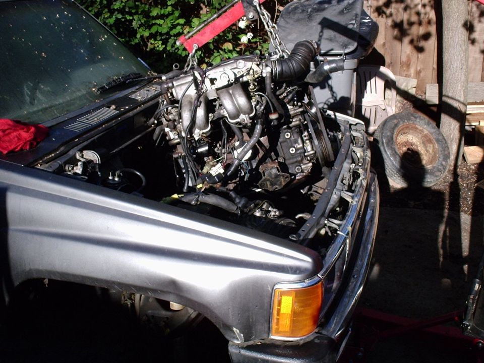 Engine suspended by chain hoist over the open engine bay of a gray Toyota pickup, hood removed