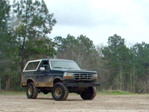 1993 Ford Bronco - A muddy Ford Bronco sits in a dirt lot with trees in the background.