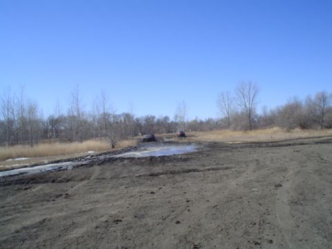 1981 Ford Bronco - Two SUVs are stuck in a muddy area with trees in the background under a clear blue sky.