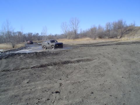 1981 Ford Bronco - Off-road vehicles navigate a challenging muddy terrain under a bright blue sky.
