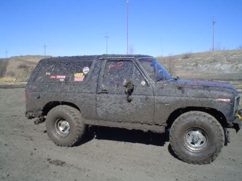 1981 Ford Bronco - Passenger-side profile of a lifted 1980s full-size Ford Bronco plastered in wet mud, sitting on tall mud-terrain tires and slotted steel wh…