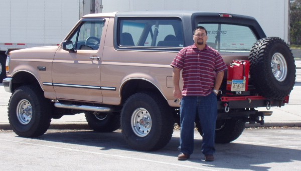 Man standing beside a lifted tan Ford Bronco with rear swing-out spare tire carrier and red fuel can