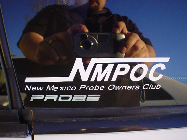 Reflection of person holding camera on car window with 'NMPOC New Mexico Probe Owners Club PROBE' sticker