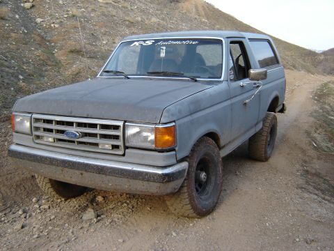 1991 Ford Bronco - A gray Ford Bronco drives on a dirt road in a hilly, arid landscape.
