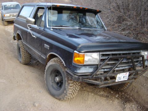 1991 Ford Bronco - A dark blue 1991 Ford Bronco is seen on a dirt road, accompanied by another vehicle in the background.