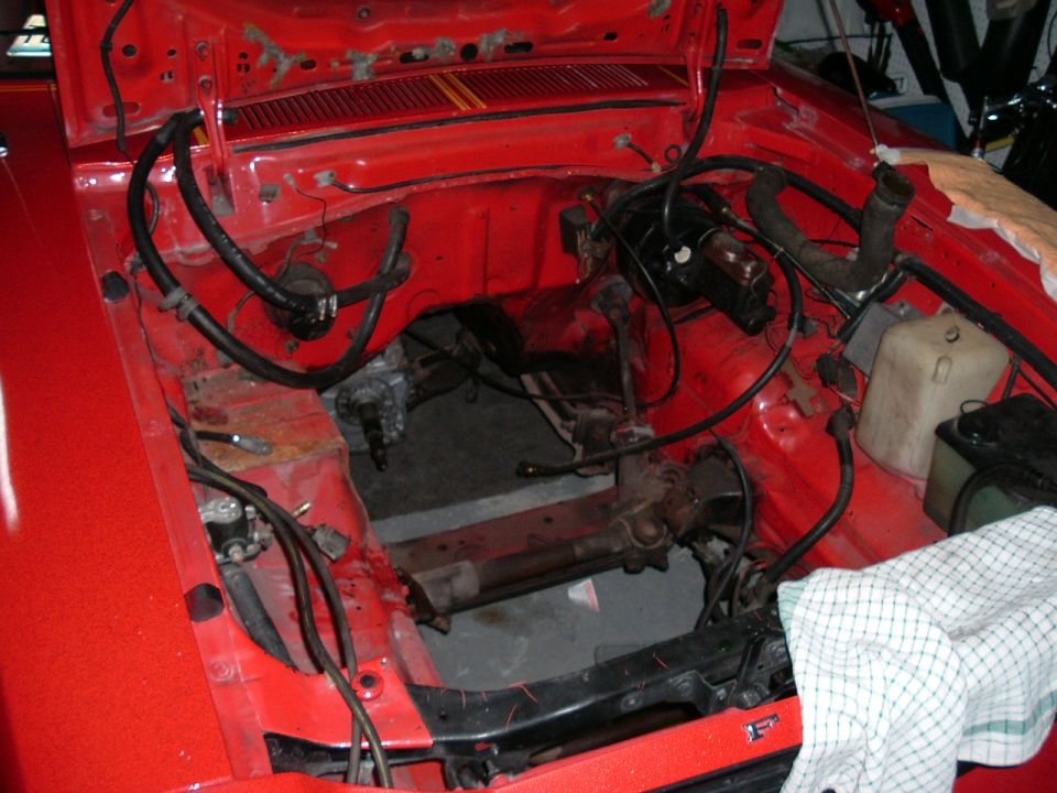 Red car engine bay with brake booster, hoses, and wiring visible. Engine is missing