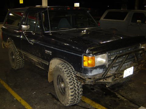 1991 Ford Bronco - Three-quarter front night shot of a mud-splattered black lifted Ford Bronco in a lit parking lot, riding on aggressive mud-terrain tires ov…
