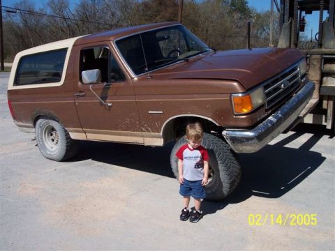 1987 Ford Bronco - A young child stands next to a lifted Ford Bronco, dwarfed by its large tires.