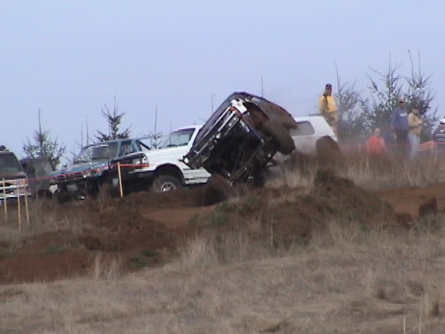 Several off-road vehicles are in a mud pit, with one truck flipped on its side