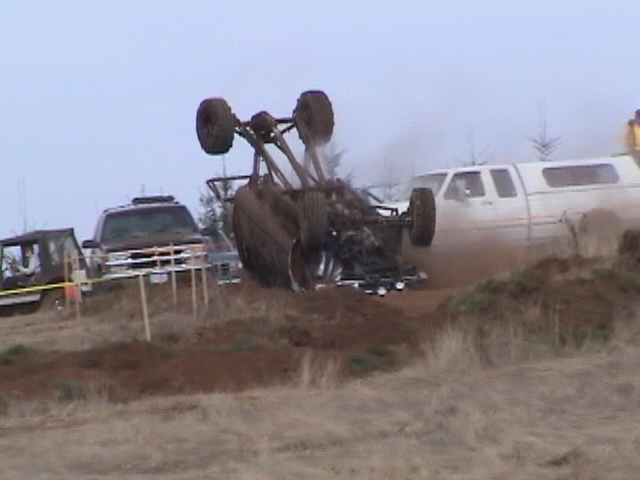 Mud-covered 4x4 flipped upside down mid-rollover on a dirt hillclimb with spectators behind a rope line